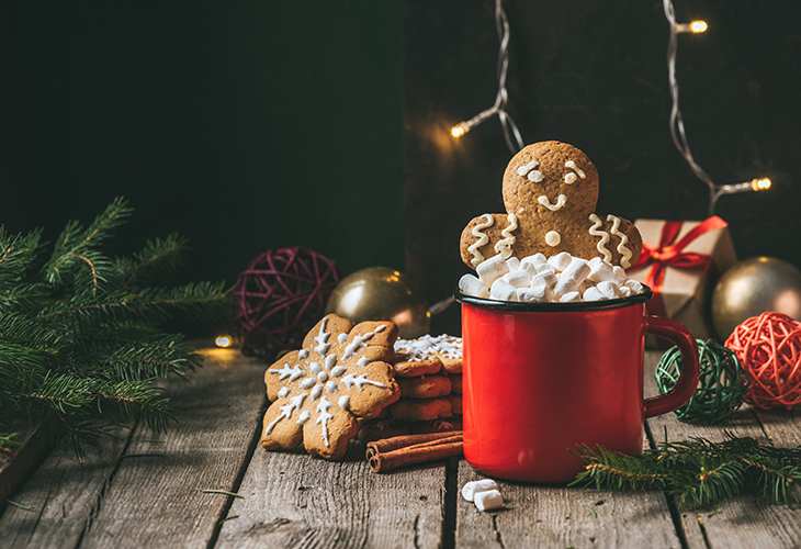 A gingerbread cookie in a mug of hot chocolate with other cookies and Christmas decorations scattered around.